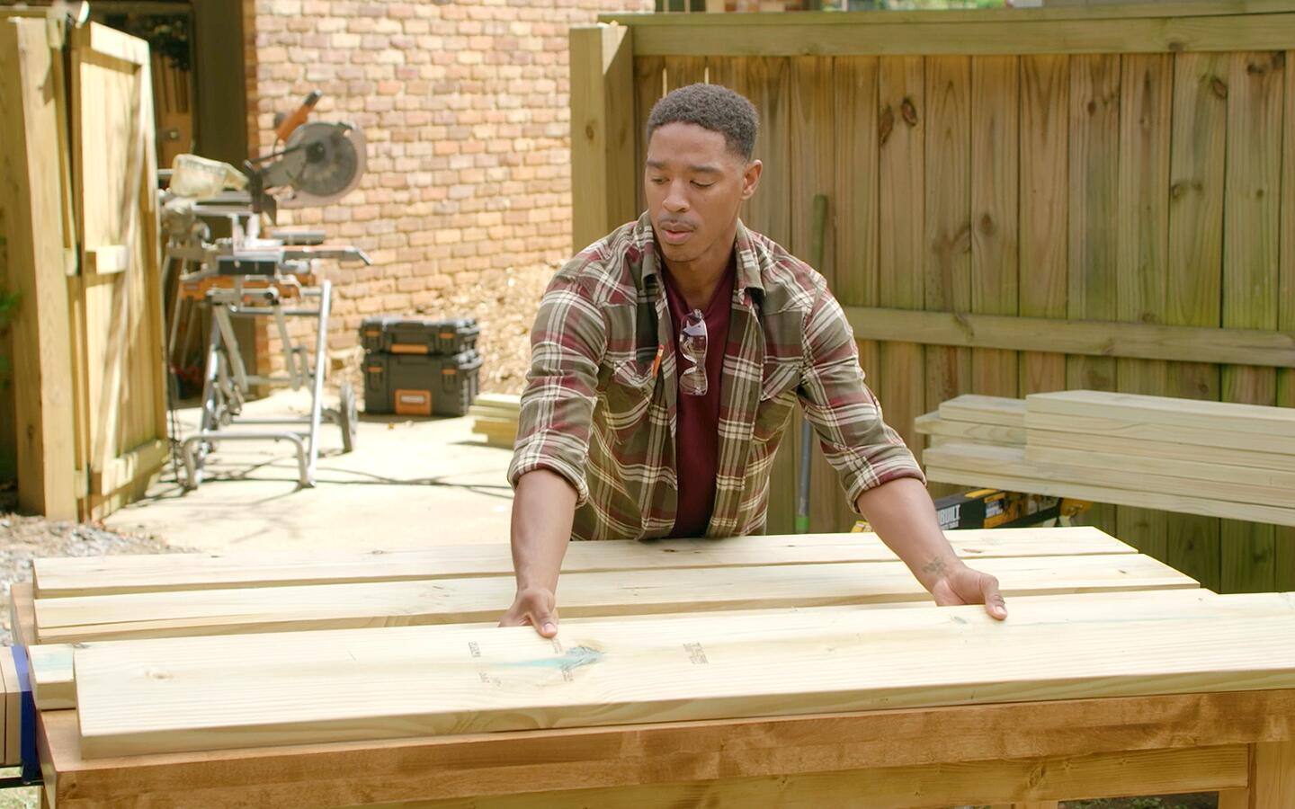 A man assembles the top of a picnic table. A wooden fence and sliding compound miter saw can be seen in the background. A man assembles the top of a picnic table. A wooden fence and sliding compound miter saw can be seen in the background.