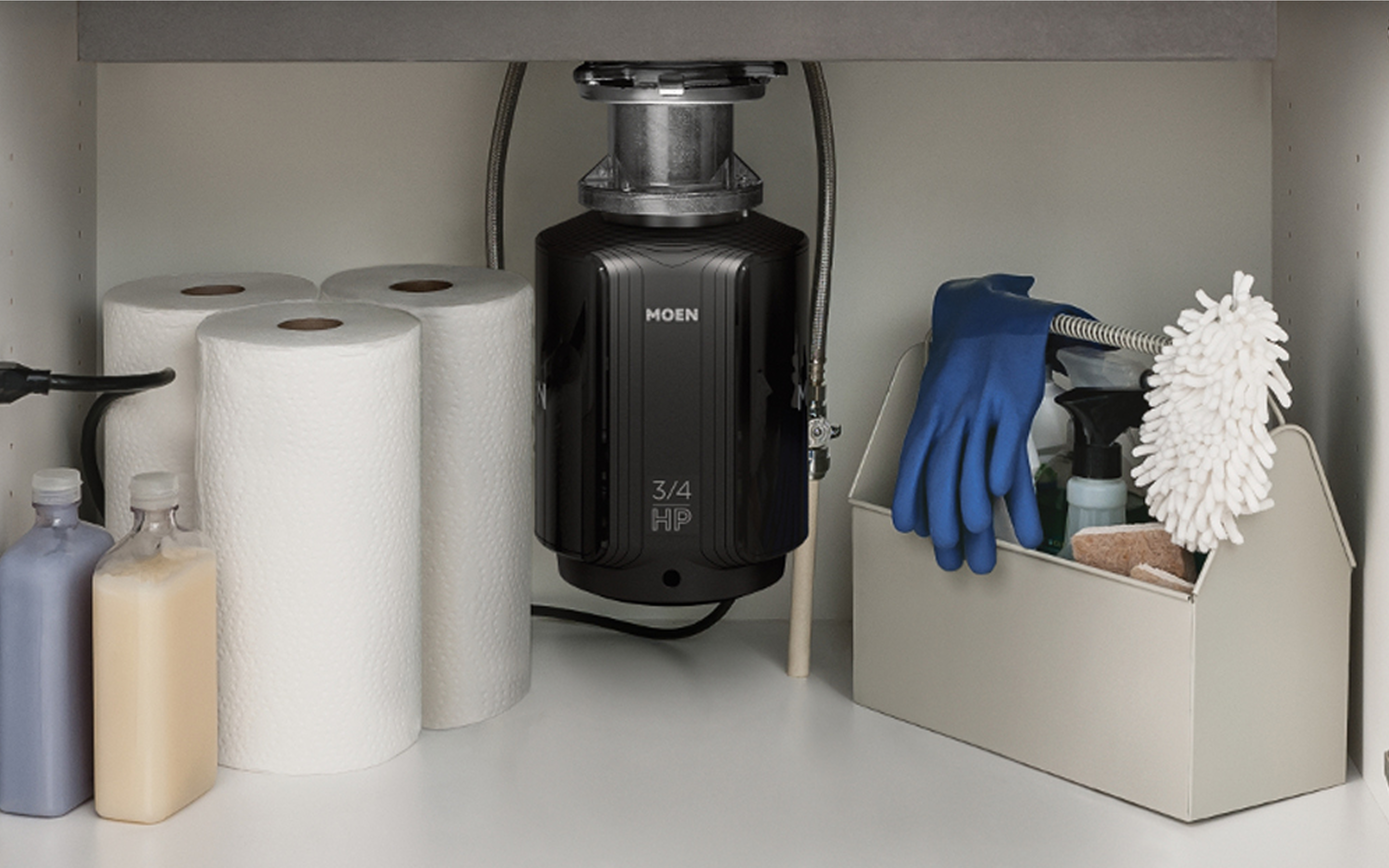 A batch feed disposal installed underneath a kitchen sink beside some paper towels and cleaning supplies.
