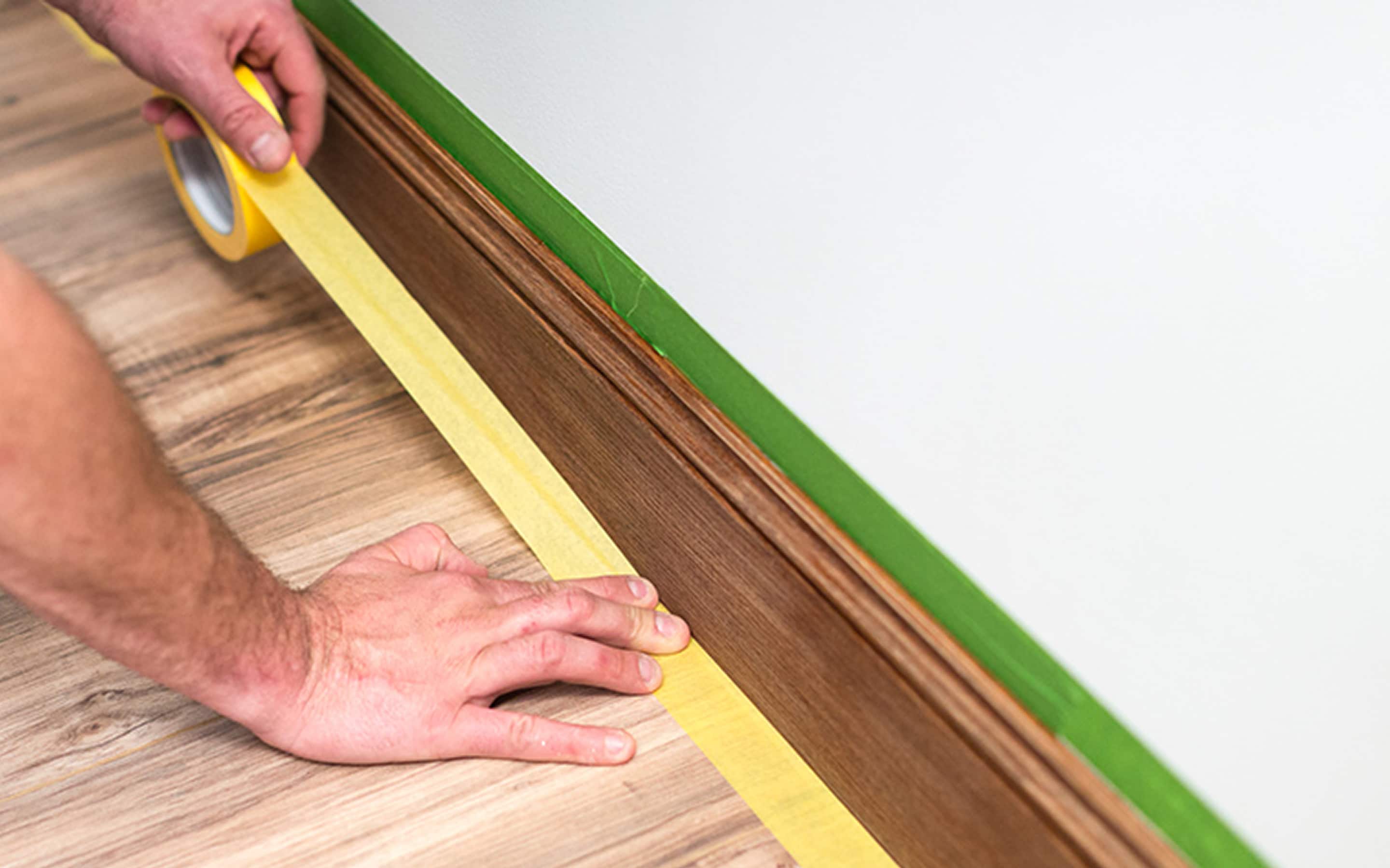 A person applies masking tape along a baseboard. A person applies masking tape along a baseboard.