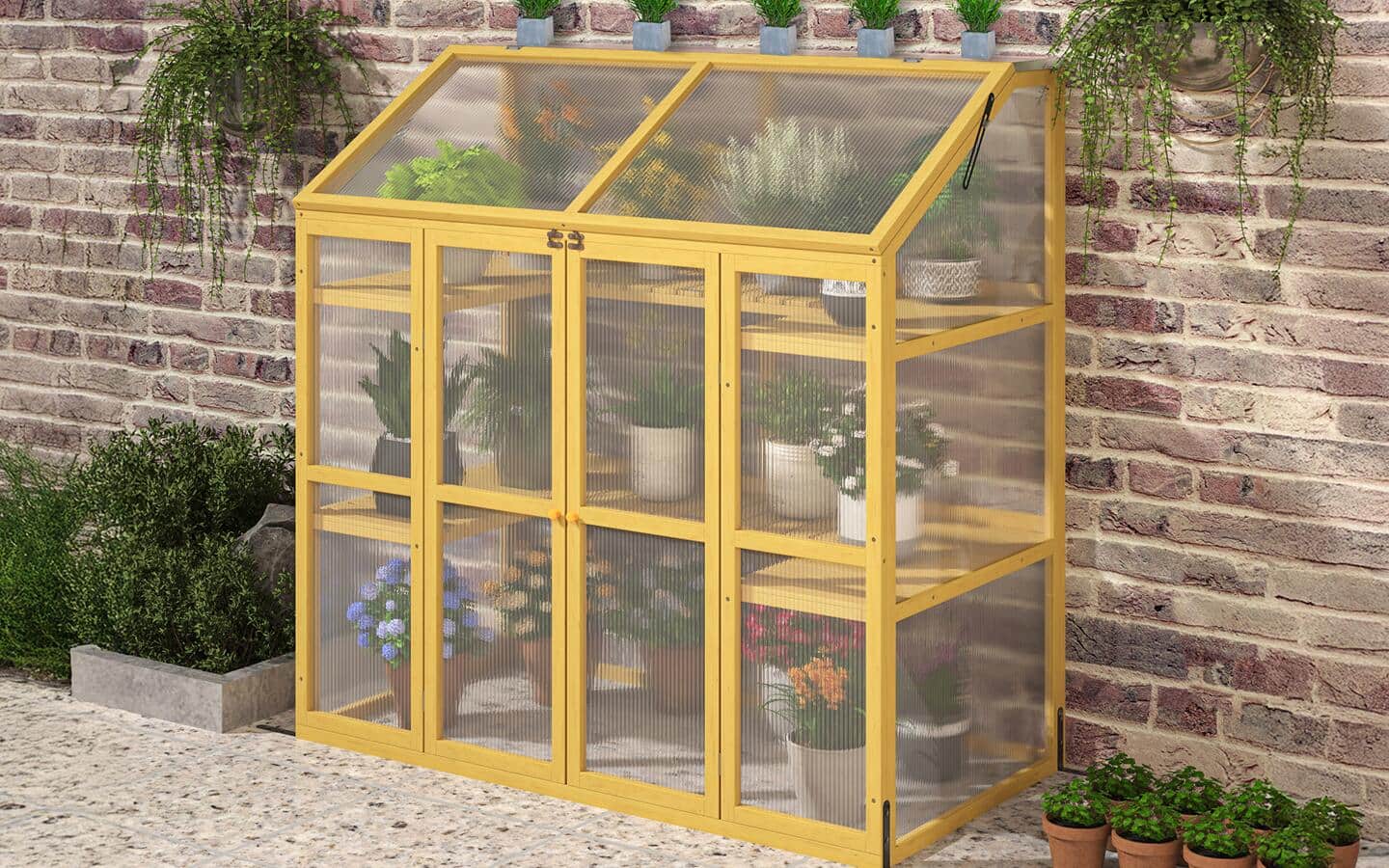A mini greenhouse with plants inside set against a brick wall in a garden