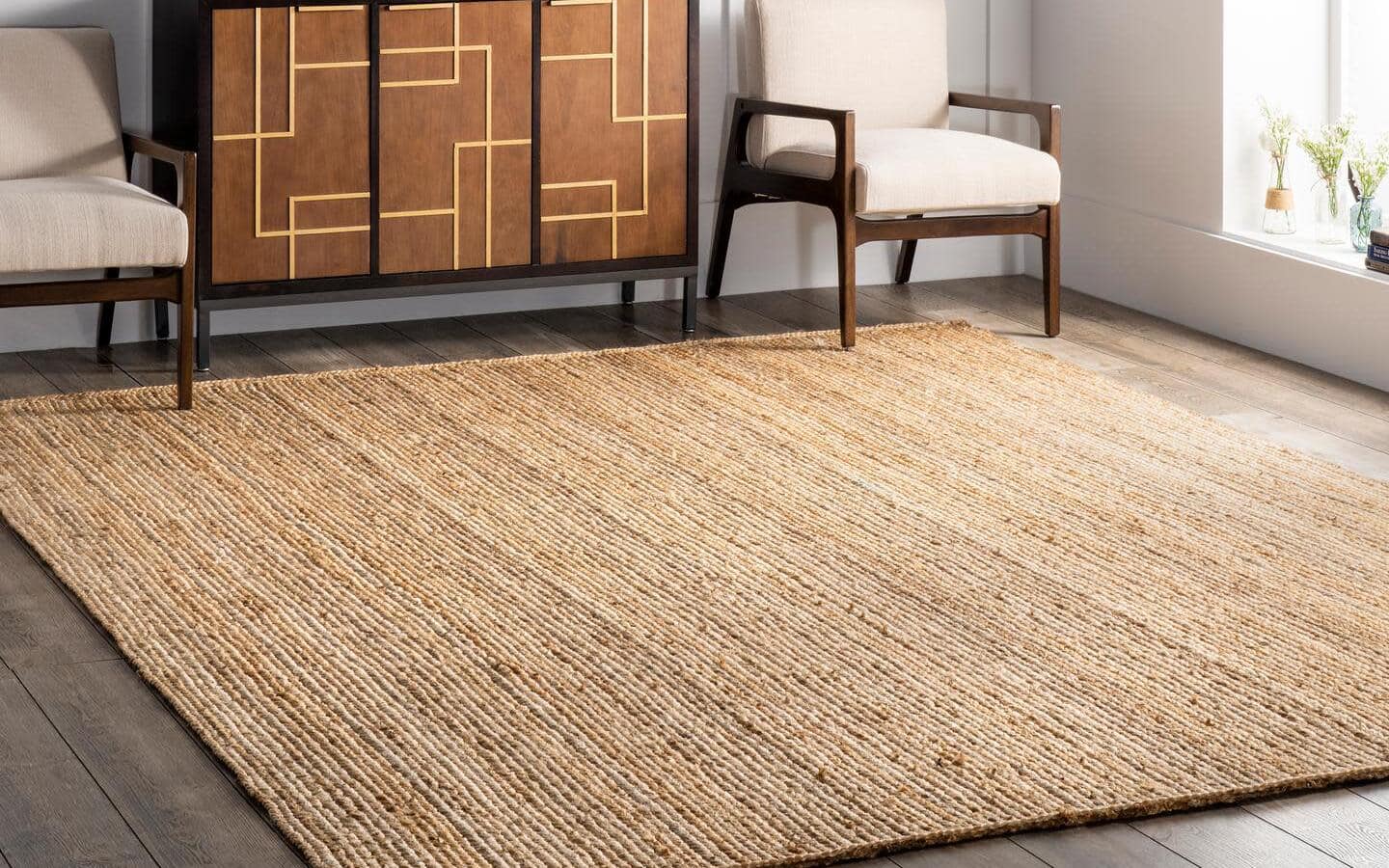 A rug made from natural materials lays next to a window, a sideboard and two chairs.