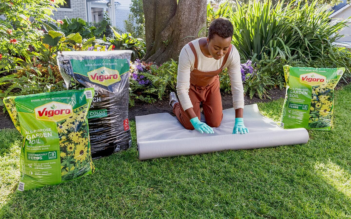 A person kneels on a lawn with a roll of landscape fabric and bags of soil and mulch