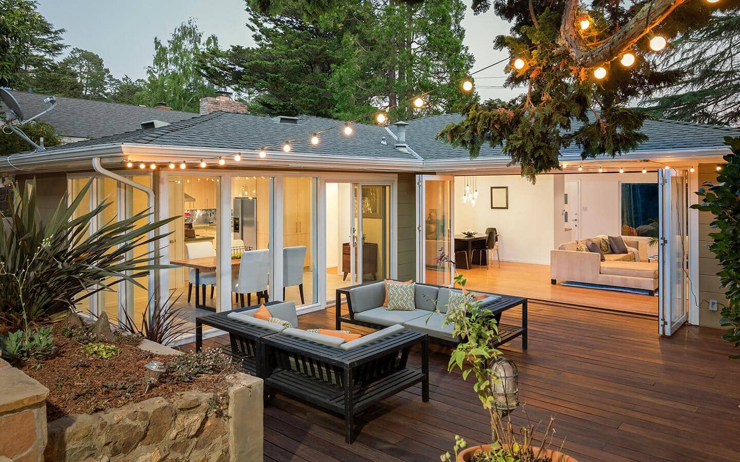 String lights hang over an attached platform deck with patio furniture and wide doors leading into a living room. String lights hang over an attached platform deck with patio furniture and wide doors leading into a living room.