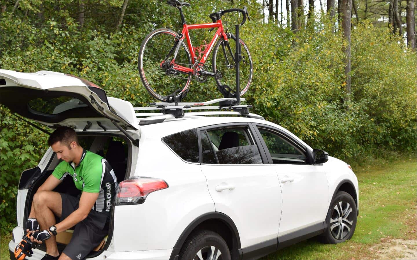 A man in the back of a car with a roof-top bike rack holding a bicycle.