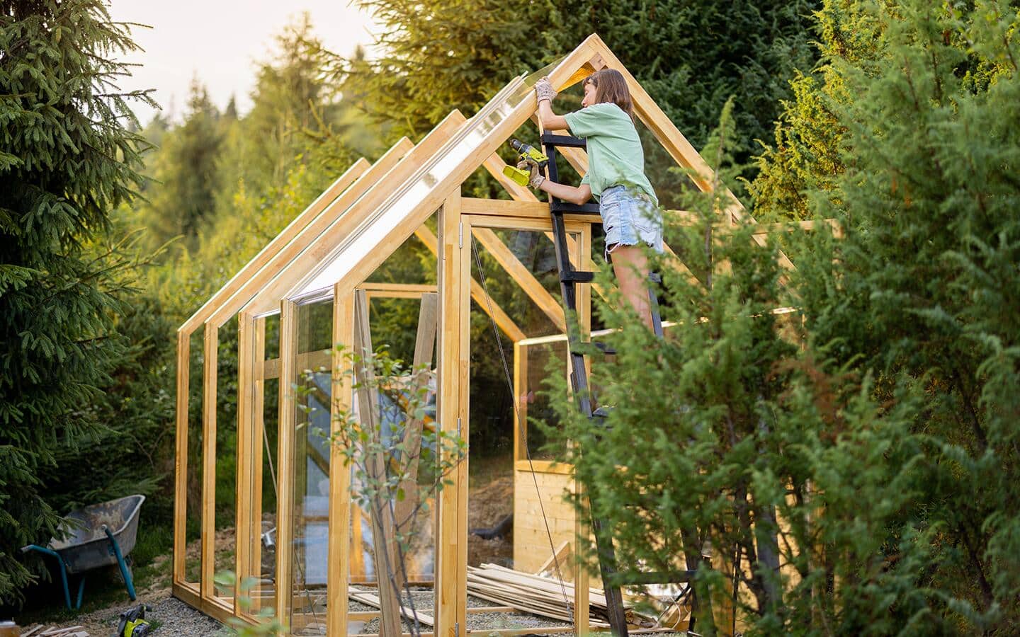 Person stands on a ladder and adds roof panels to a greenhouse structure