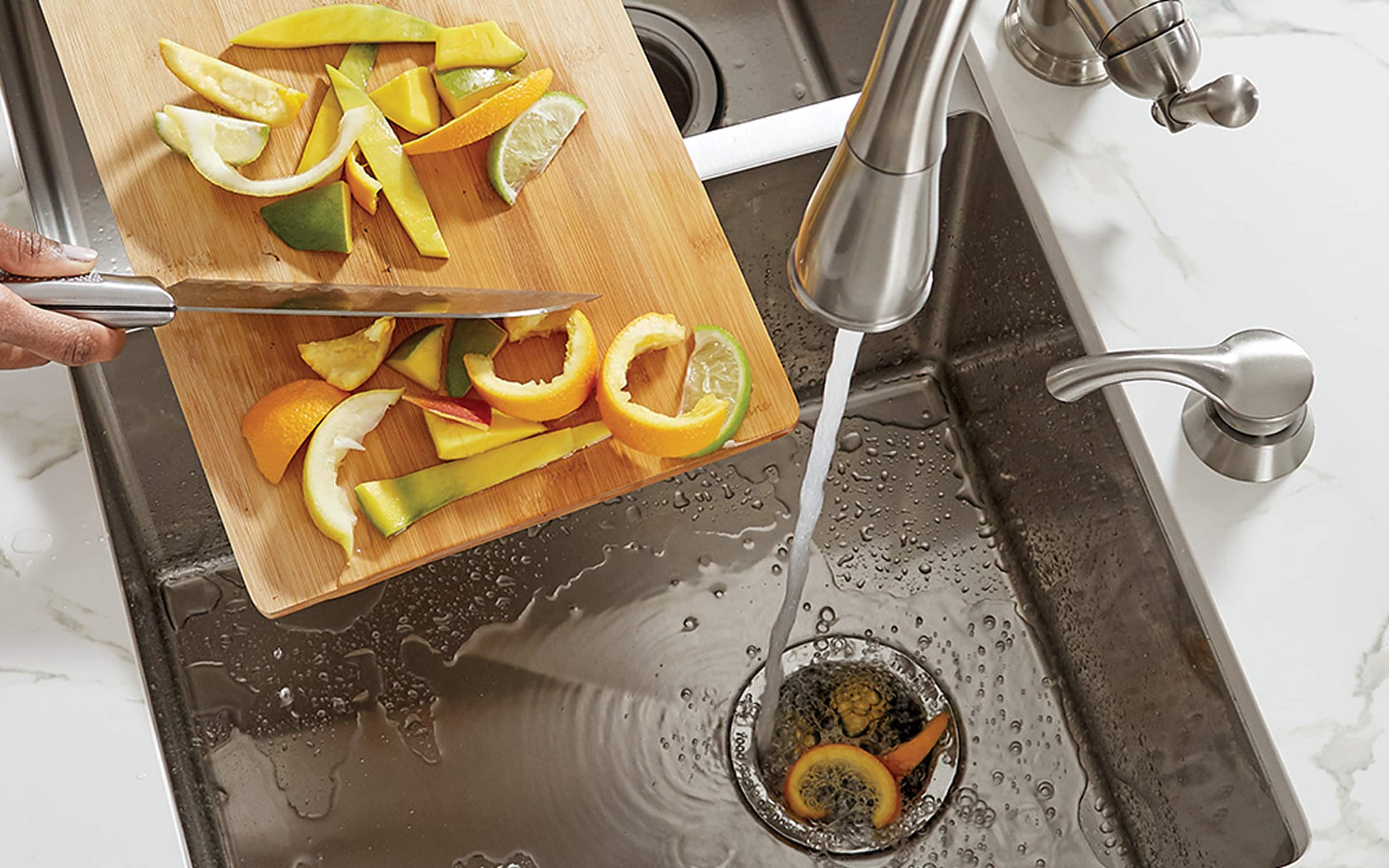 Someone using a knife to scrap citrus peelings off a cutting board into the opening of a garbage disposal while water is running.