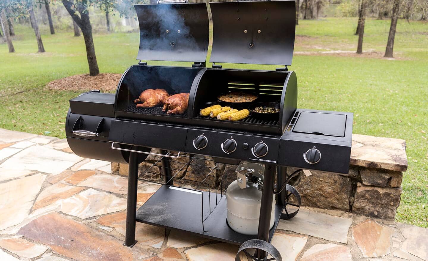 Meat, corn on the cob and other food cooks in a combination grill-smoker on a stone patio.