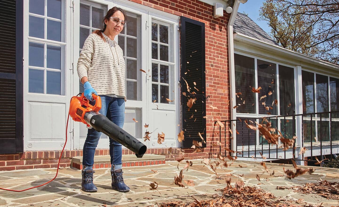 A person uses a corded leaf blower to clear leaves from a patio.