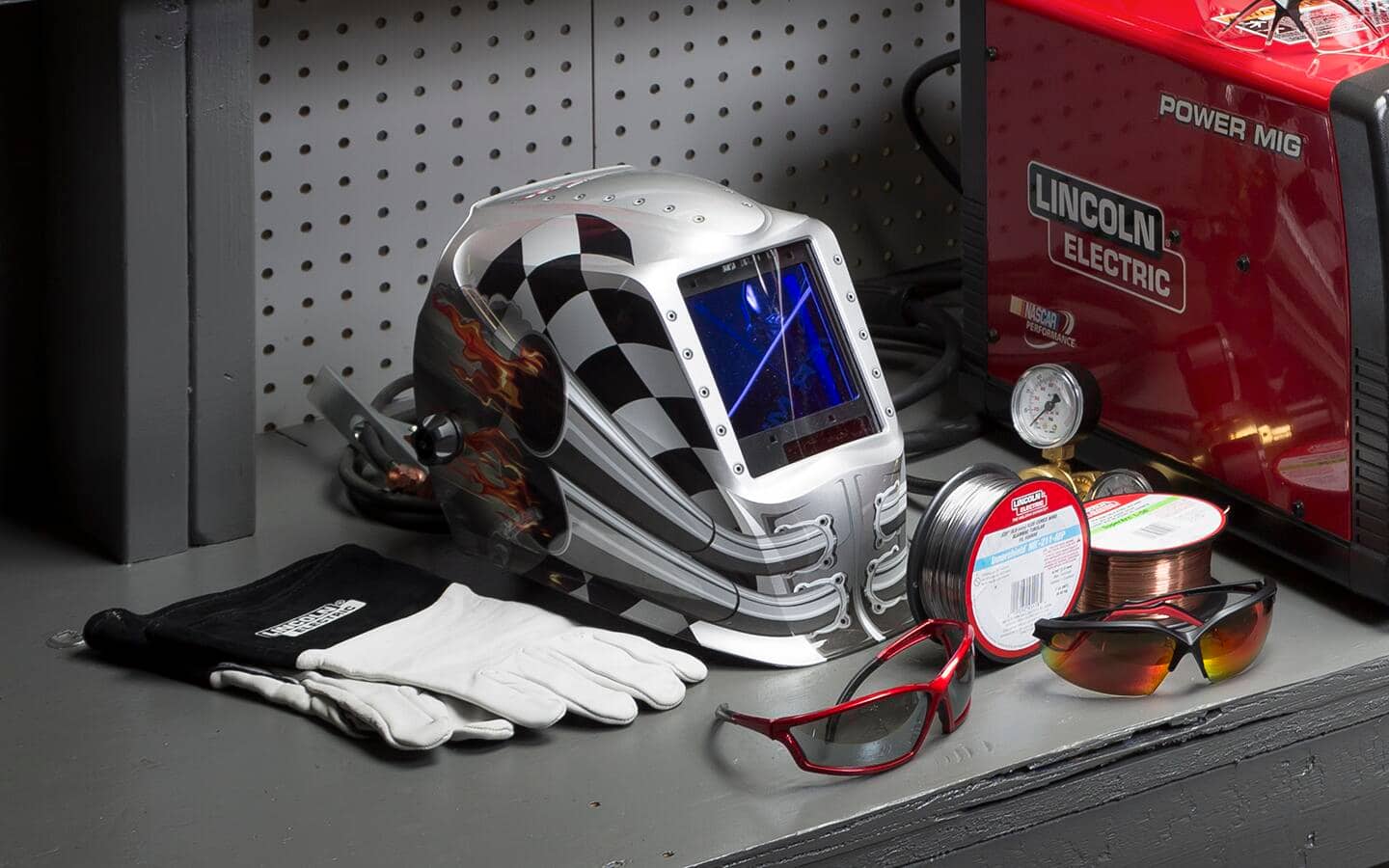 Welding equipment, including a helmet, gloves and safety glasses, on a shelf.