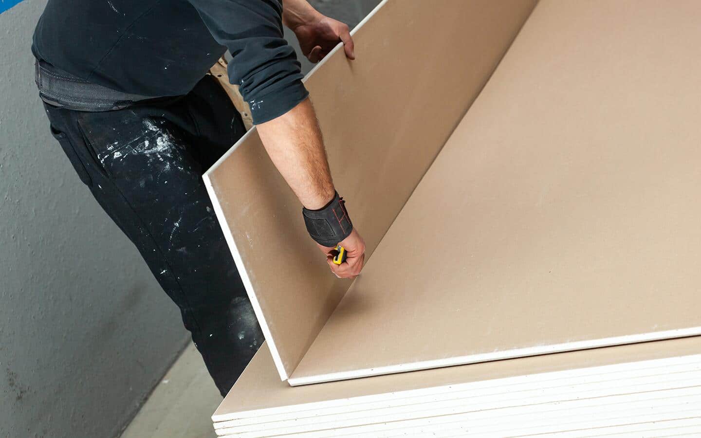 An individual in black clothes uses a utility knife to cut a piece of drywall. An individual in black clothes uses a utility knife to cut a piece of drywall.