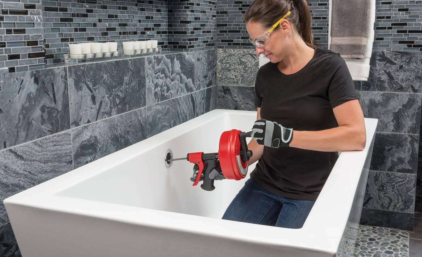 A woman uses a drain auger through the overflow drain of a bathtub.