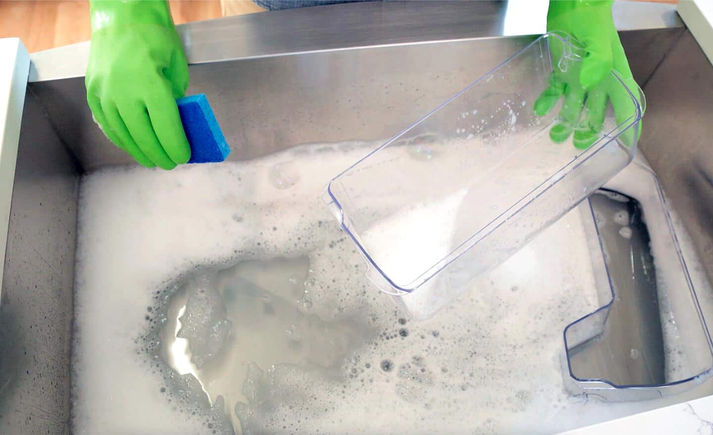 A person wearing gloves washes a removable refrigerator container in a sink filled with soapy water.