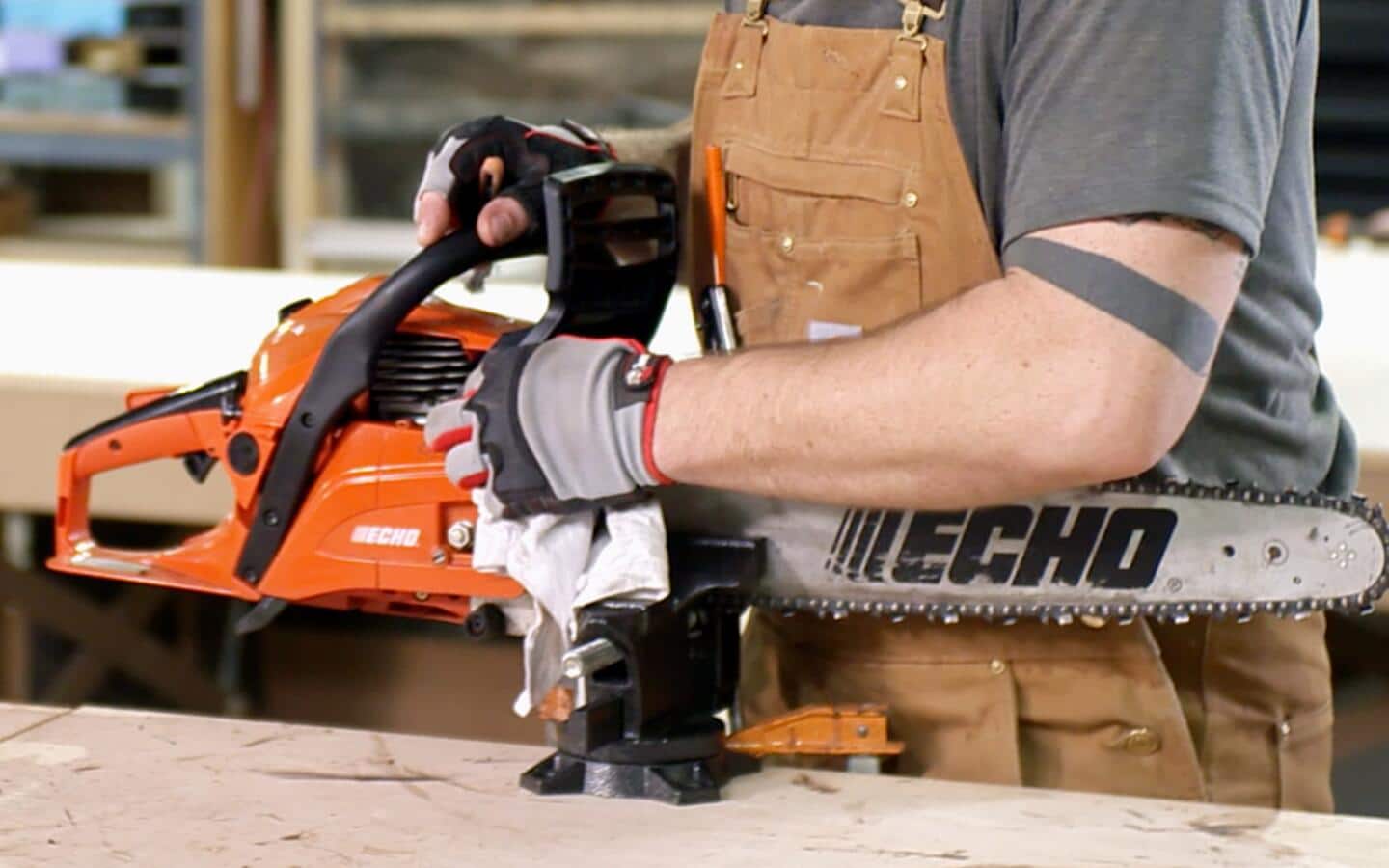 A person wearing brown coveralls uses a rag to clean a chainsaw secured to a workbench with a bench vise. A person wearing brown coveralls uses a rag to clean a chainsaw secured to a workbench with a bench vise.