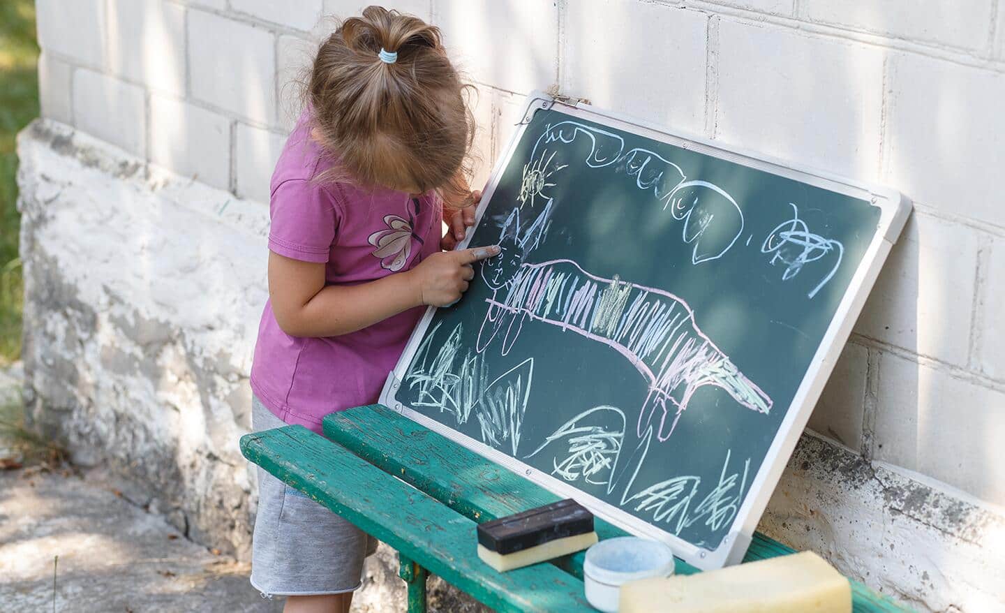A child drawing on a chalkboard outside on a green bench.