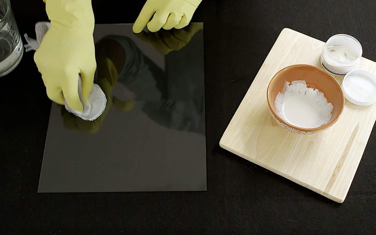 A gloved person applies glass polish prepared in a bowl to a glass pane's scratches.