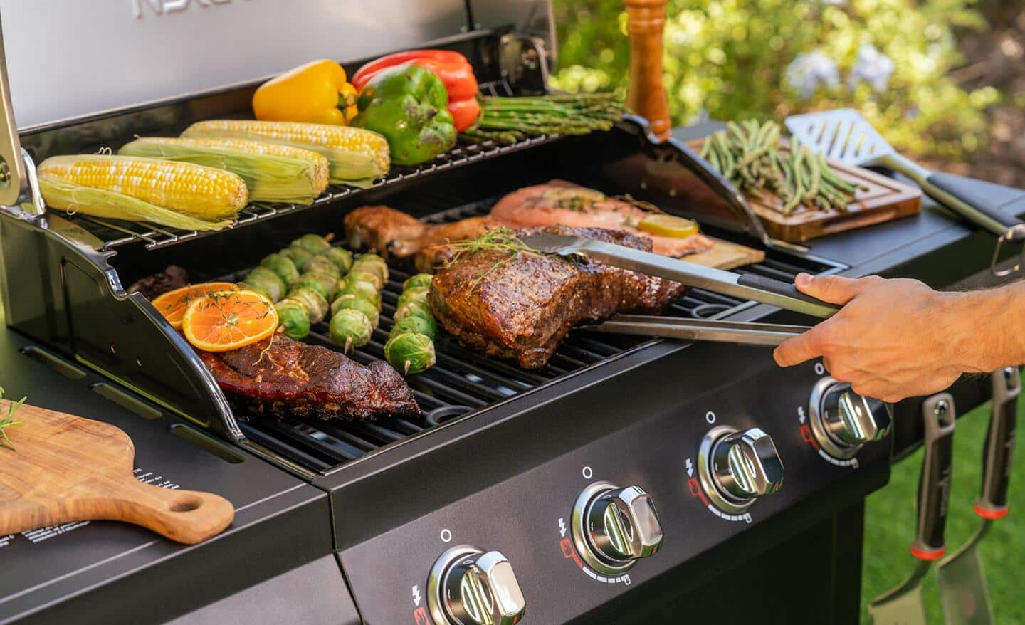 A person tending to a variety of food cooking on a gas grill.