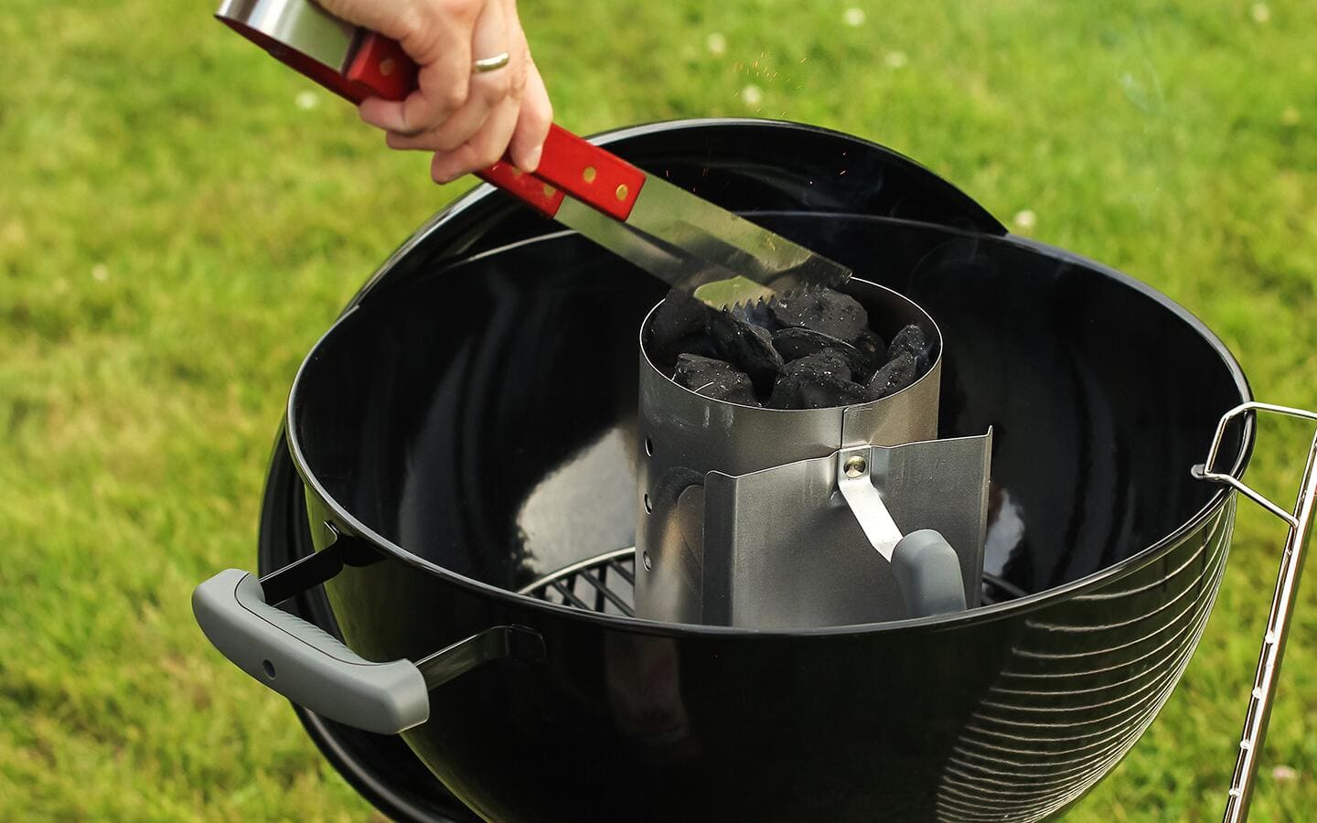 A person adds coals to a charcoal chimney sitting inside a charcoal grill. A person adds coals to a charcoal chimney sitting inside a charcoal grill.