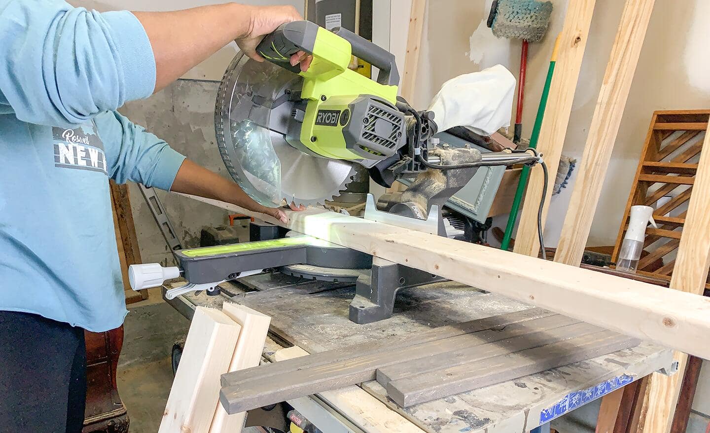 A person braces a board on a miter saw with one hand as he pushes the saw handle forward to make the cut.