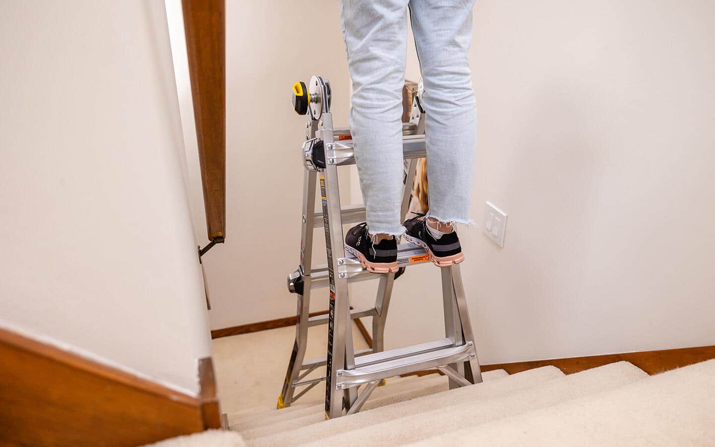 A person stands on a multi-position ladder that is set up in a stairway.