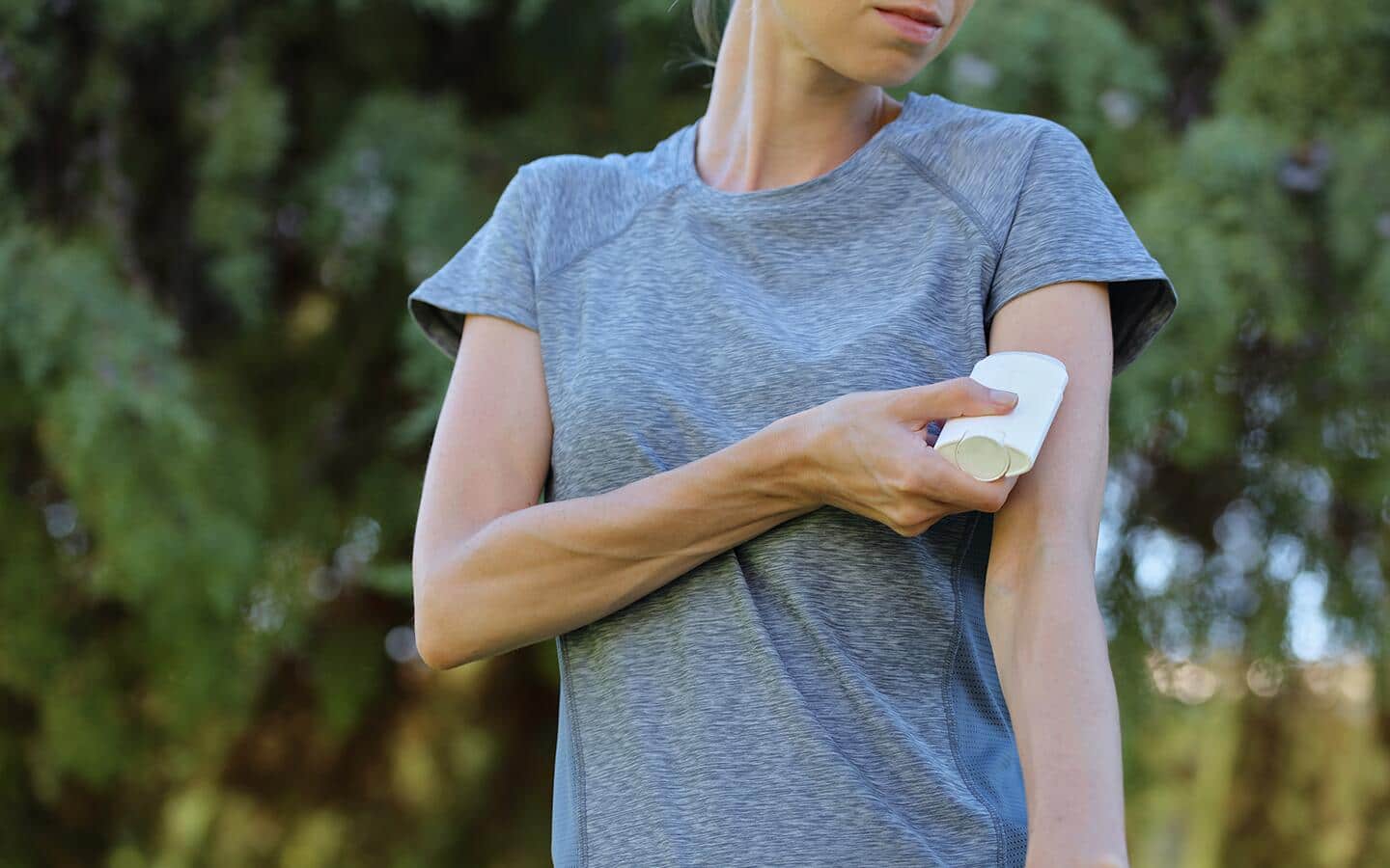 A woman wearing a gray T-shirt applies roll-on insect repellent to her arm.