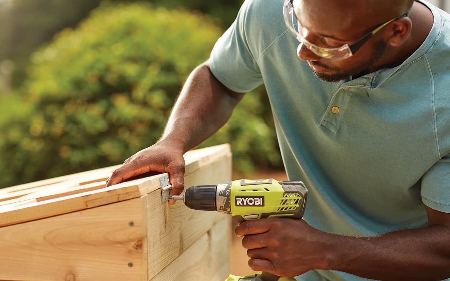 A person uses a drill to drive screws into the hinges that attach the top of cold frame to the base. 