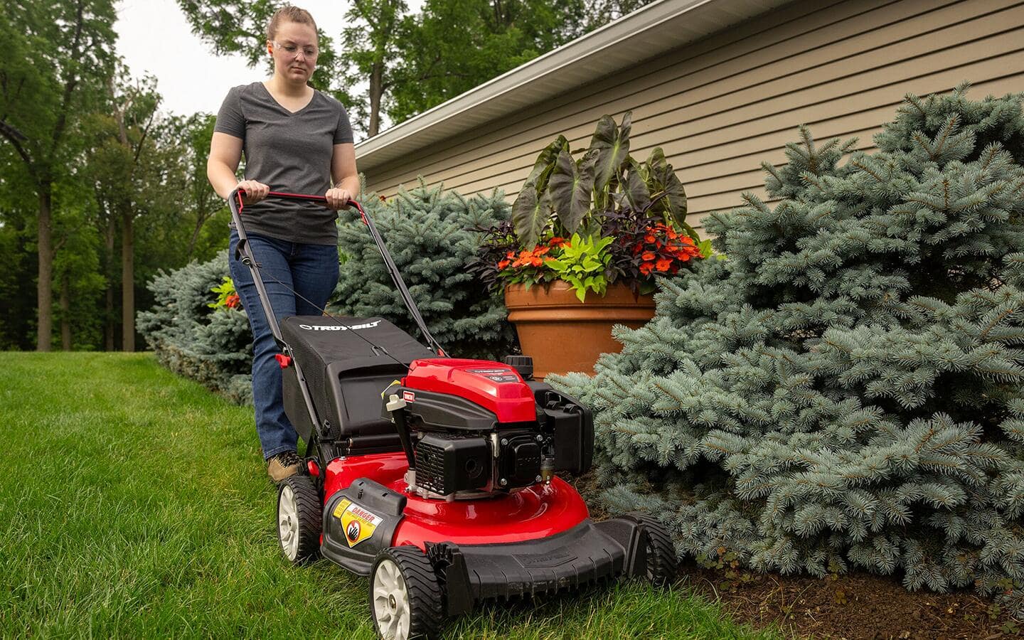 An individual uses a red gas push mower in a yard alongside shrubs. An individual uses a red gas push mower in a yard alongside shrubs.
