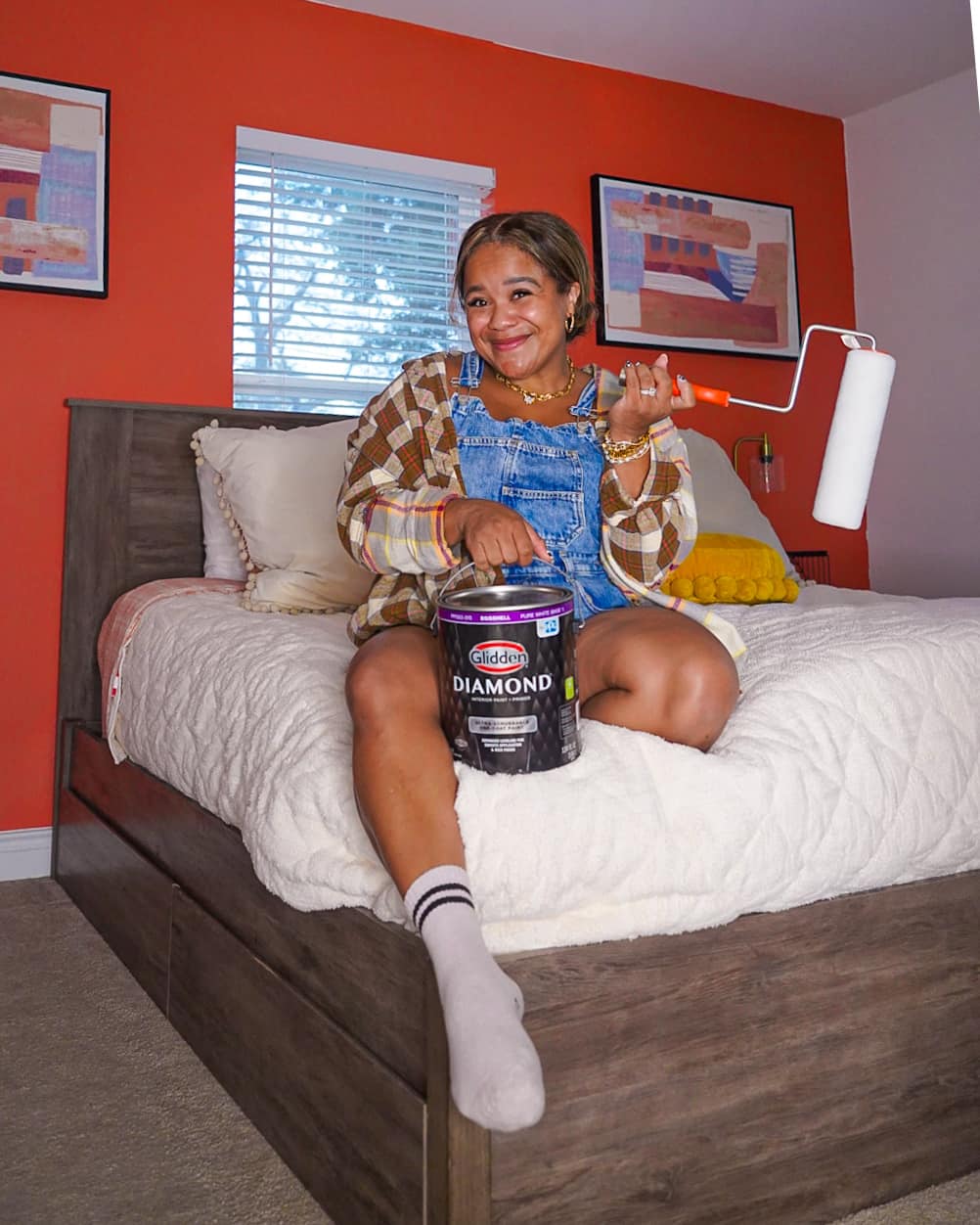 A person sitting on a bed holding a paint can and paint roller.