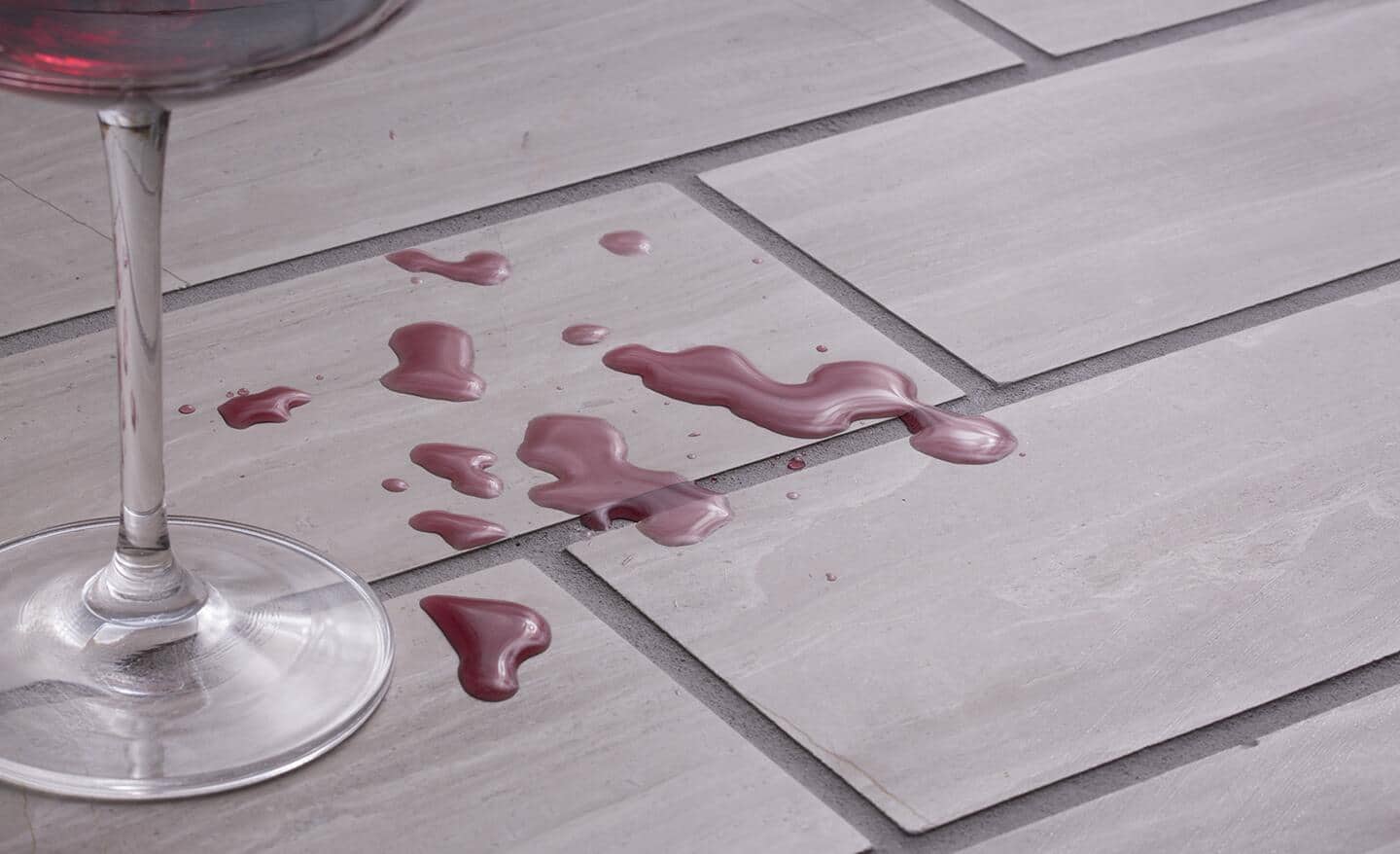 A tiled floor with grout has a glass with spilled red liquid on the tile's surface.