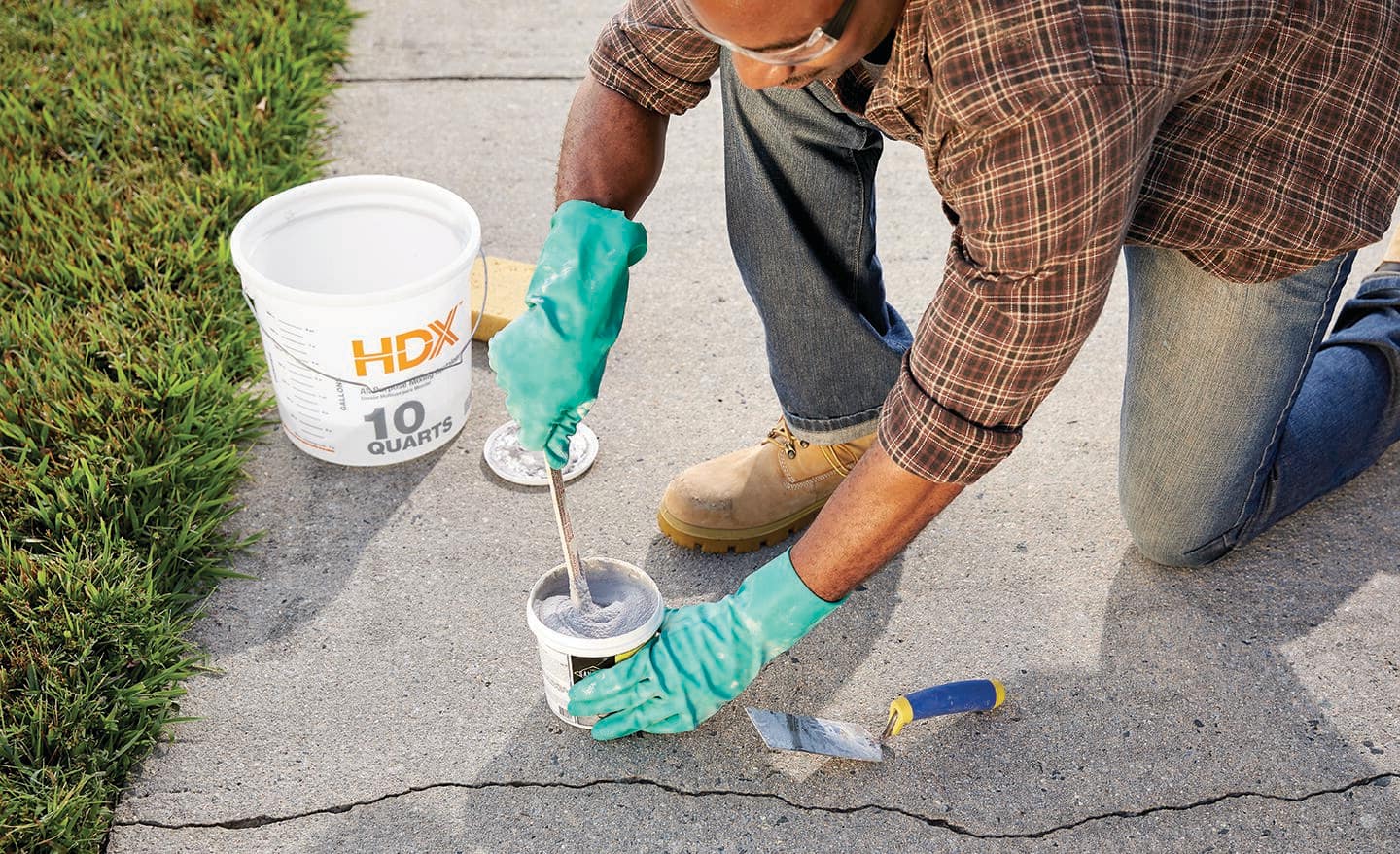 A person stirs pre-mix concrete patch repair while kneeling next to a crack in a concrete sidewalk.