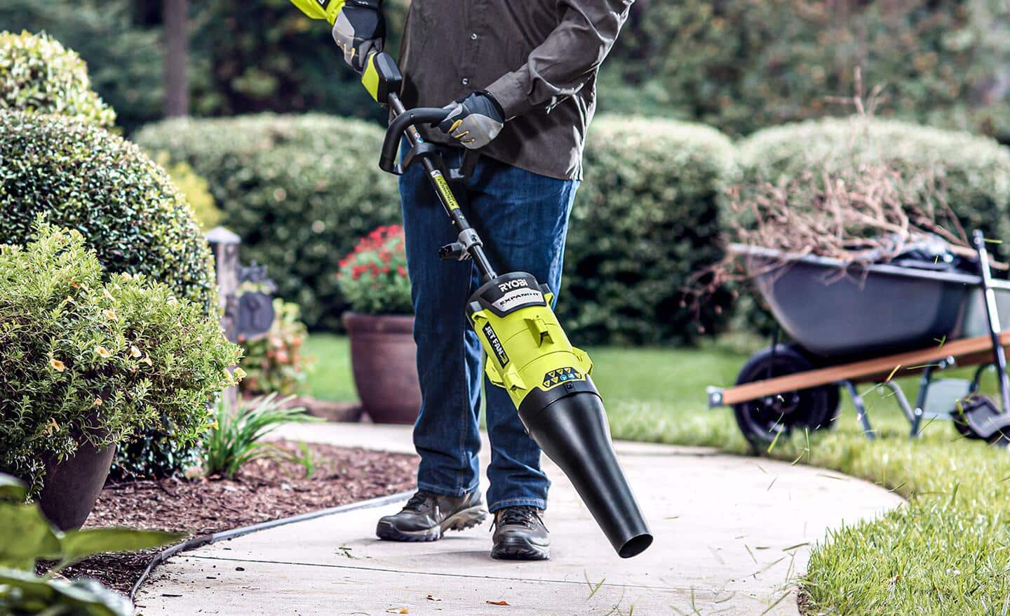 A person uses a string trimmer with a leaf blower attachment to remove leaves and debris from a paved path.