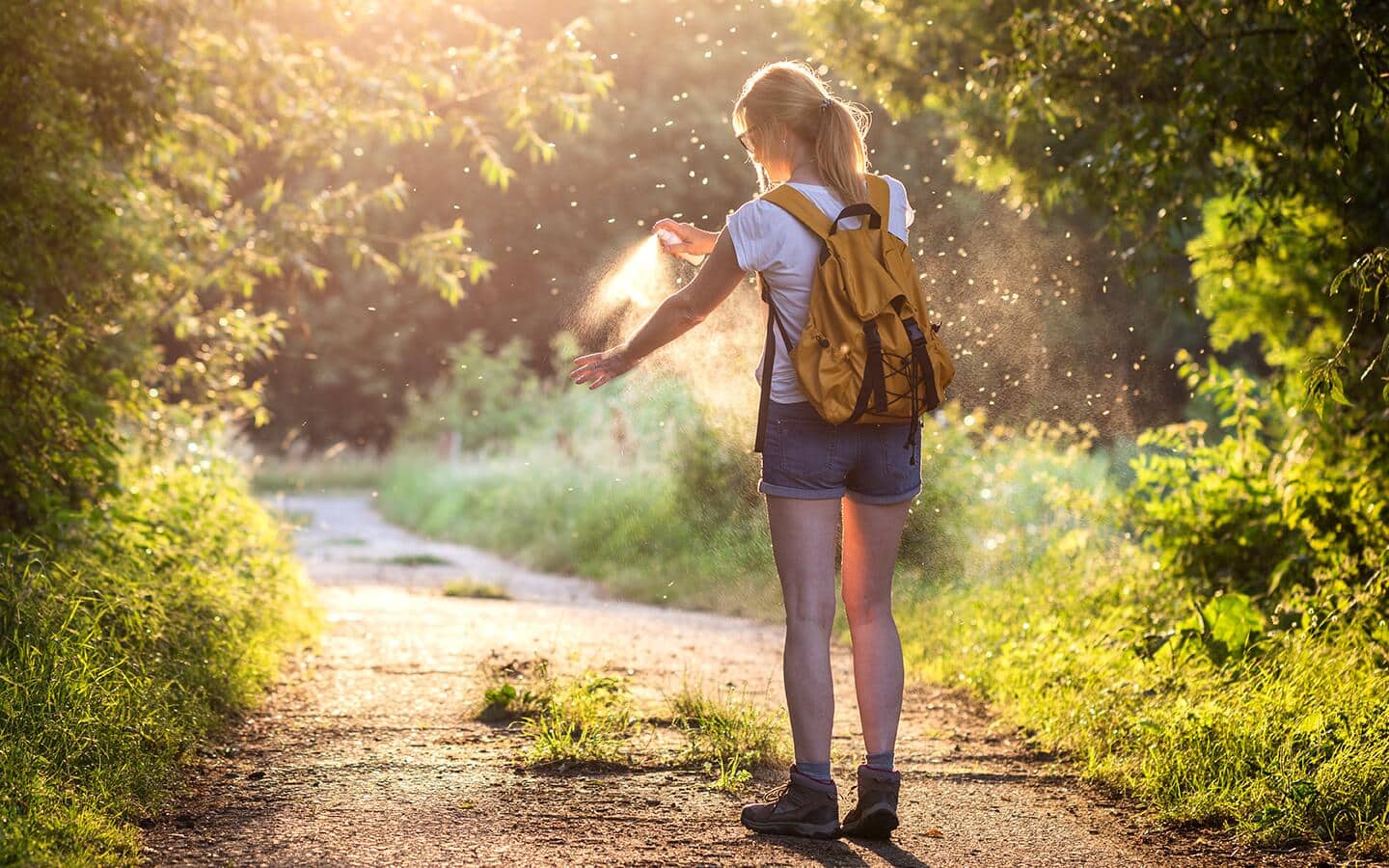 A woman with a backpack stands on a sunlit hiking trail and sprays insect repellent on her arm.