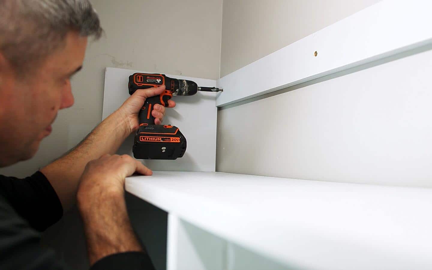 A person screwing in a support for a top shelf with a cordless drill.
