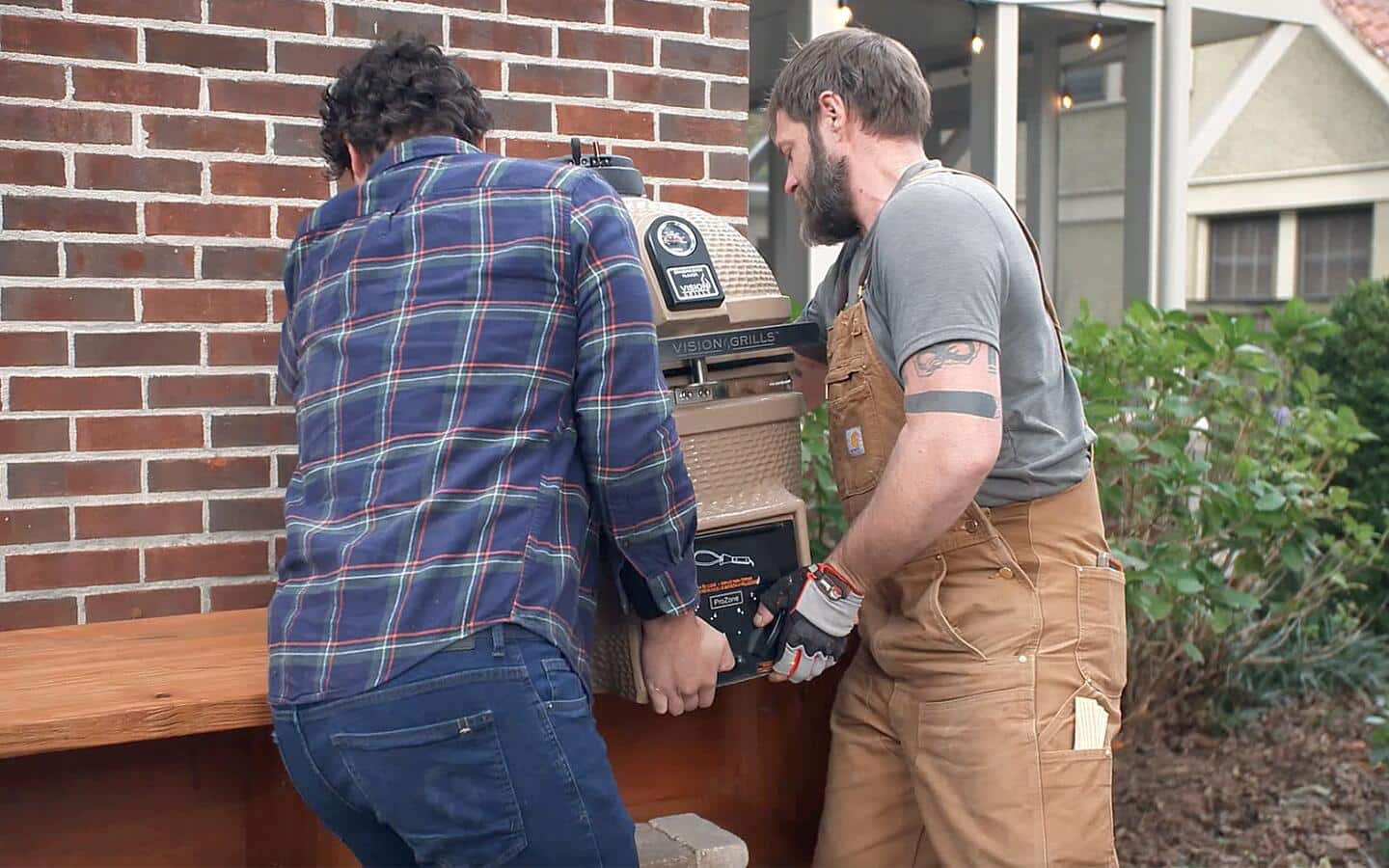 Two people lift a small Kamado grill into place beside a home's side wall.