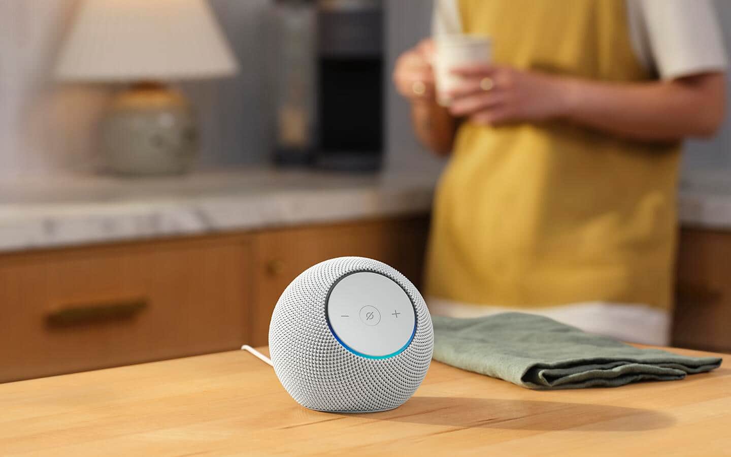 A smart speaker on a kitchen countertop next to a green towel while a person holds a coffee cup in the background. A smart speaker on a kitchen countertop next to a green towel while a person holds a coffee cup in the background.