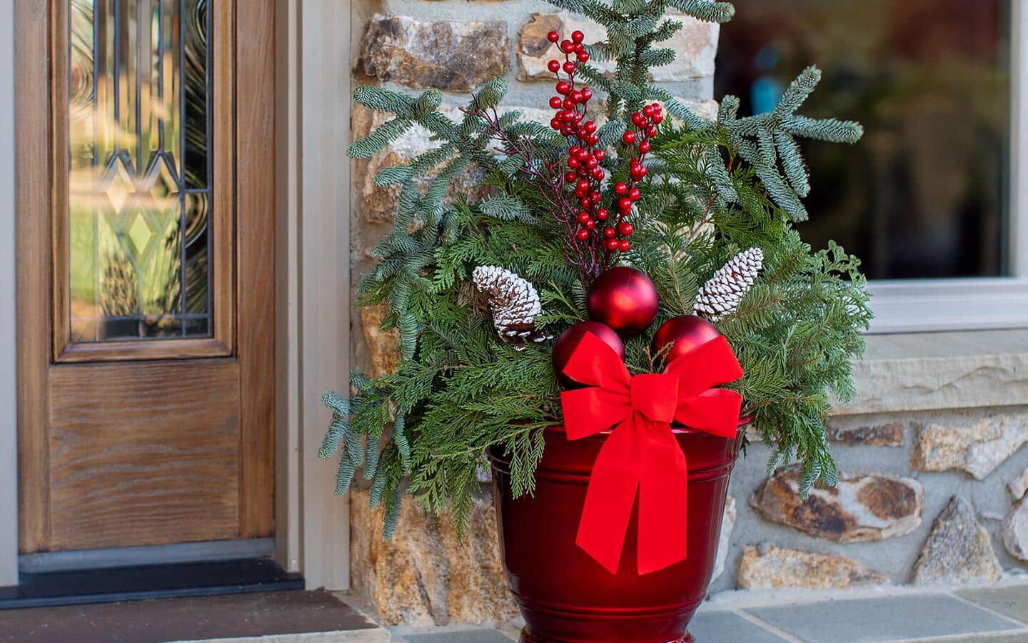 A red pot filled with evergreen branches and sprigs of red berries on a front porch.
