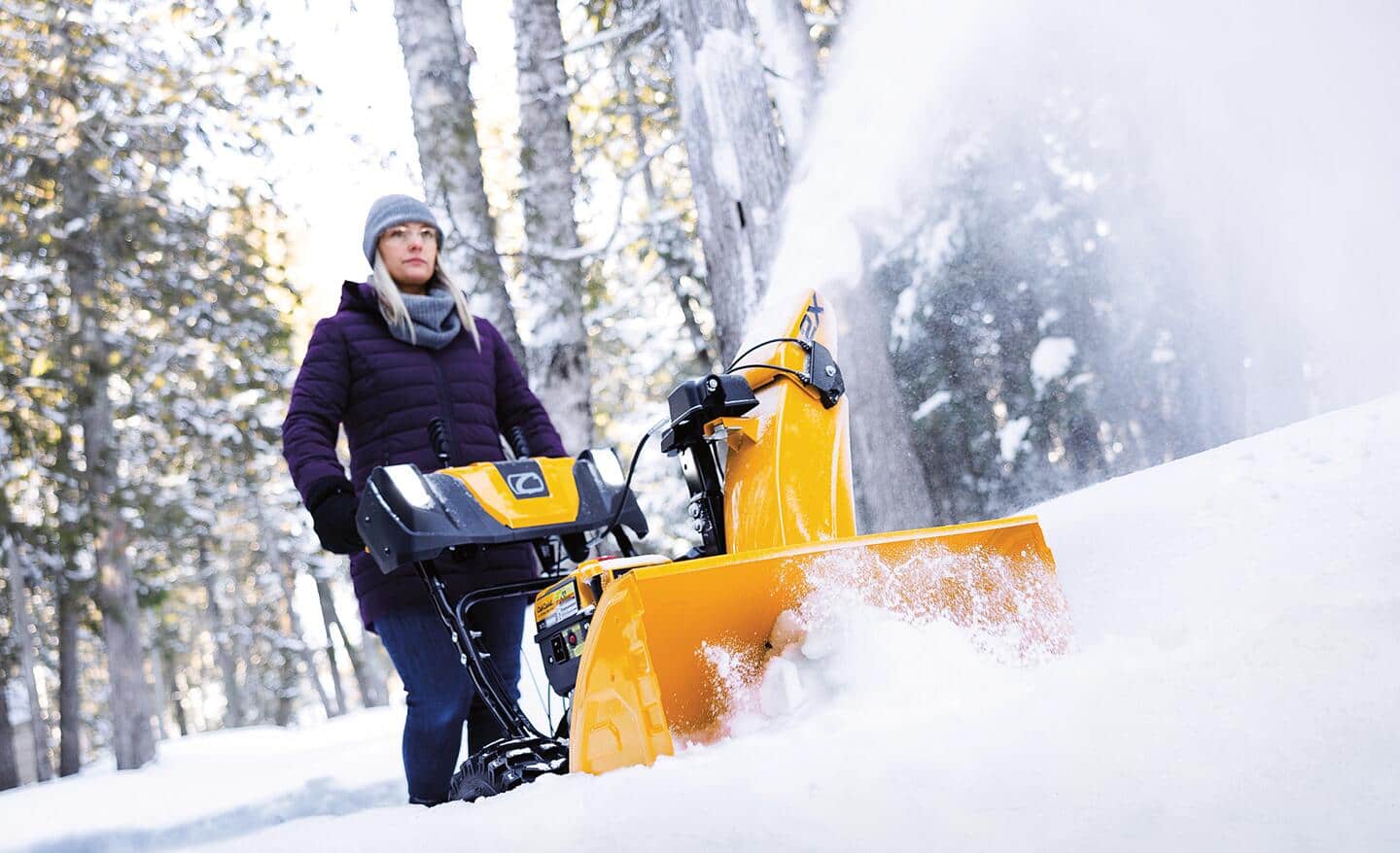 A person using a yellow two-stage snow blower in the woods.