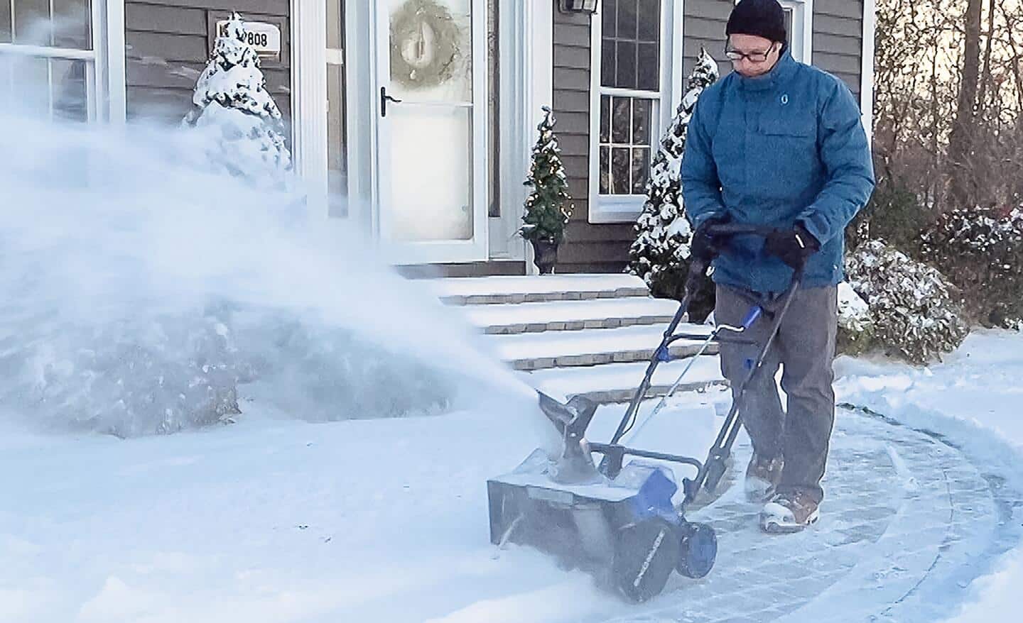 A blue cordless snow blower clearing a path in front of a house.