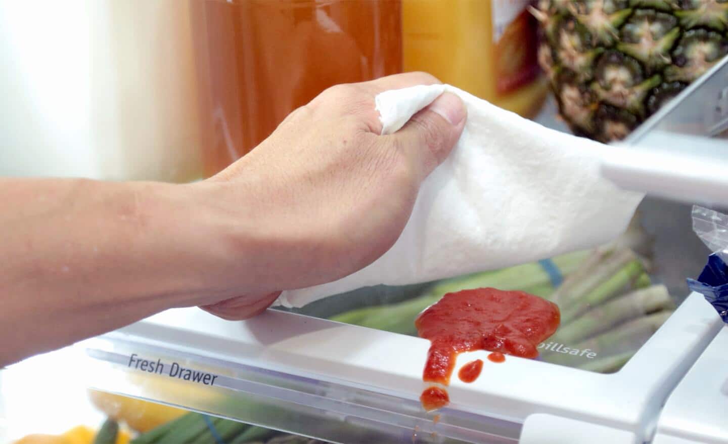 A person uses a cloth to wipe up a spill on an interior refrigerator shelf.