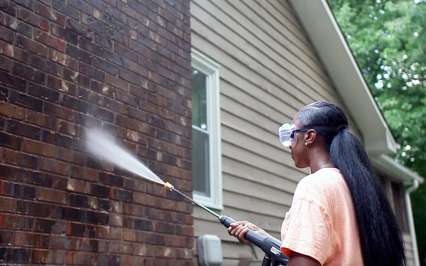 A person wearing eye goggles sprays the exterior brick wall of a house.