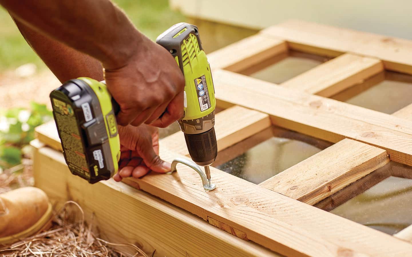 A person uses a drill to drive screws to attach a handle to the top of a cold frame. 