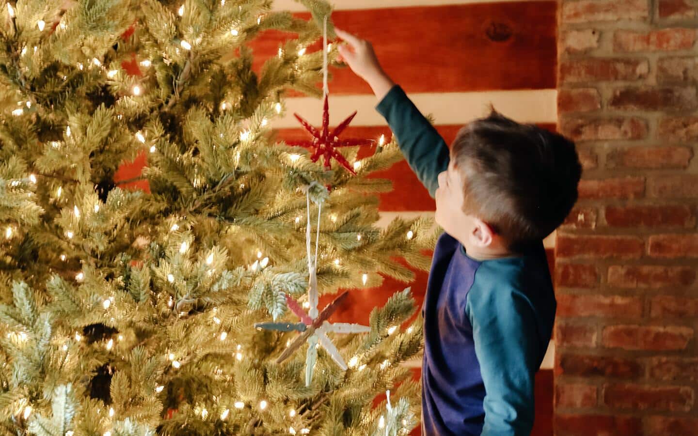 A little boy hanging red stars on a lit Christmas tree.