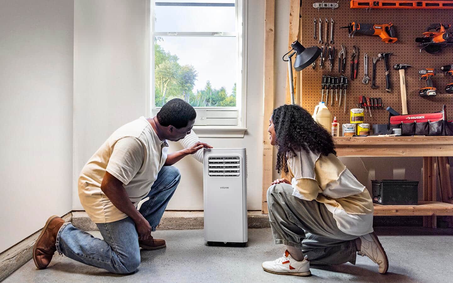 Two people look at a dehumidifier on the floor next to a work bench. Two people look at a dehumidifier on the floor next to a work bench.