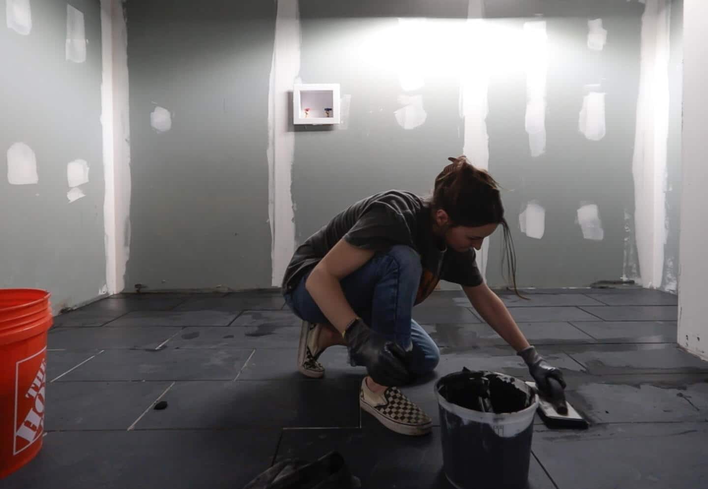 Girl in jeans and checkered shows smoothing black grout over black tiles in large basement.