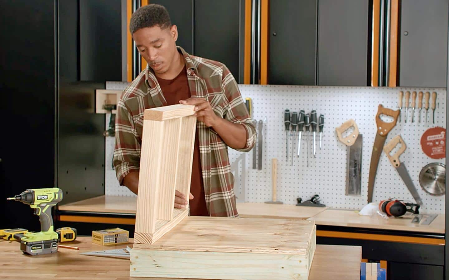 A person in a checkered shirt standing up a dog house frame's walls.