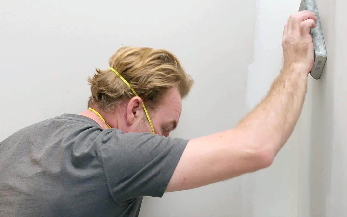 A person wearing a safety mask sands drywall with a sanding block. 