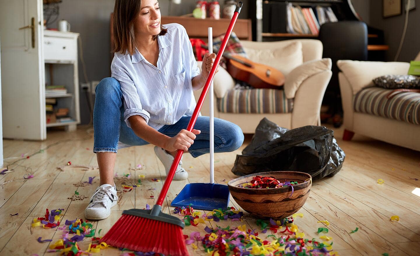 A woman using a broom and dust pan to clean up after a party.