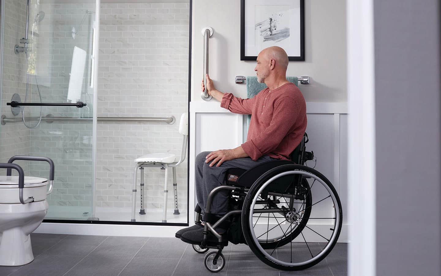A person in a wheelchair holds a grab bar in an accessible bathroom. A person in a wheelchair holds a grab bar in an accessible bathroom.