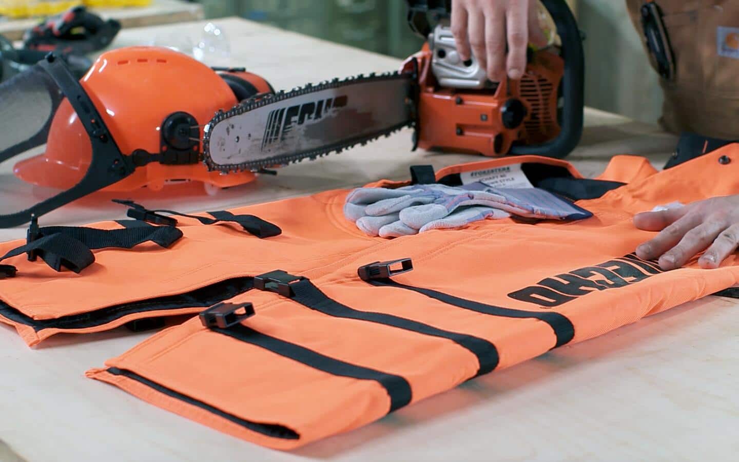 Orange safety chaps, work gloves, an orange helmet with a safety mask lie on a workbench next to a chainsaw. Orange safety chaps, work gloves, an orange helmet with a safety mask lie on a workbench next to a chainsaw.