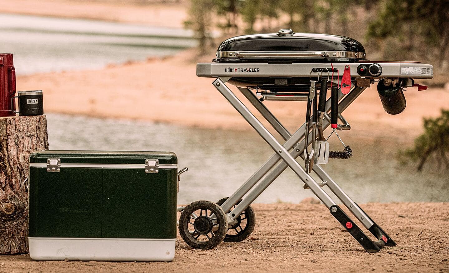 A portable grill stands next to a cooler and a tree stump.