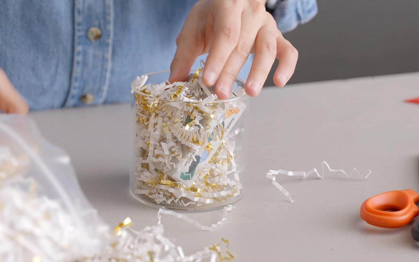 A person places a gift in a glass canister filled with shredded paper.